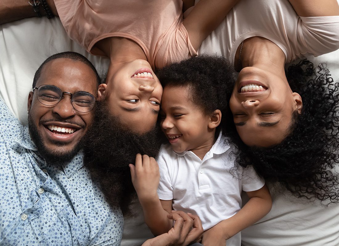 About Our Agency - Portrait Looking Down at a Cheerful Family with Two Kids Laying Down on the Bed While Having Fun Spending Time Together