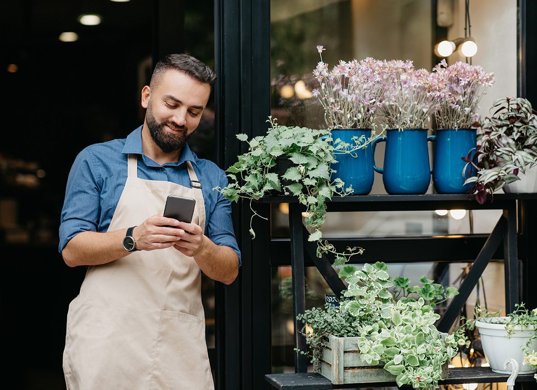 Business Insurance - Portrait of a Young Male Business Owner Wearing an Apron Standing Outside his Shop Next to Decorative Plants While Using a Phone