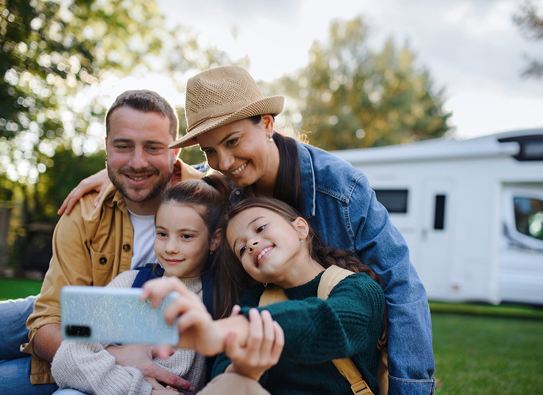 Insurance Solutions - Portrait of Cheerful Mother and Father with Two Daughters Sitting on Green Grass Taking a Selfie Photo While on a Camping Trip with an RV