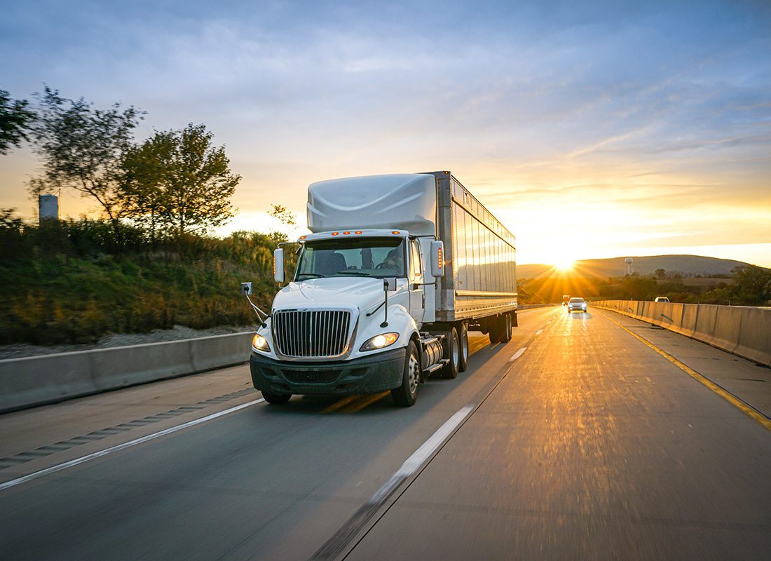 Insurance by Industry - View of a White Tractor Trailer Truck Driving Down a Scenic Highway with Trees and Mountains in the Background at Sunset